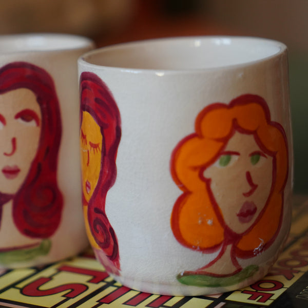 Two ceramic mugs with artistic depictions of women's faces on a table.